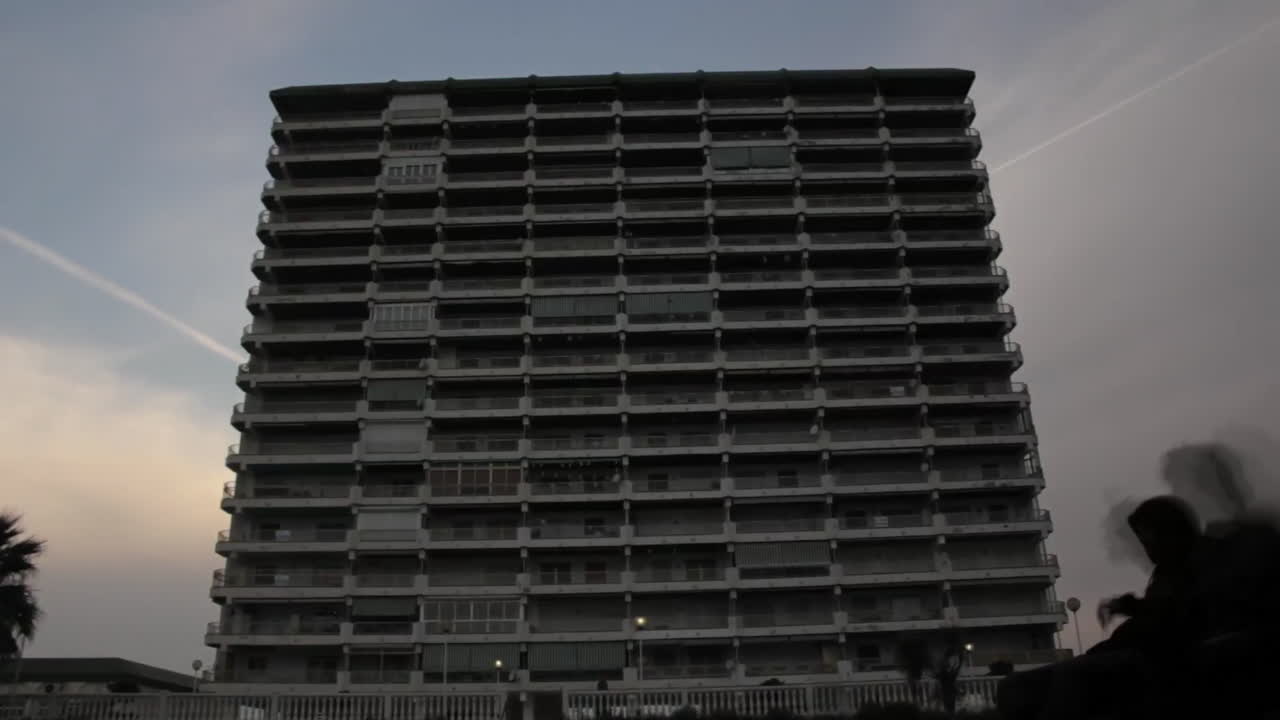 Ghostlike figures drift past a massive apartment block at dusk, blending memory and reality—evoking nostalgia, presence, and the layers of urban life