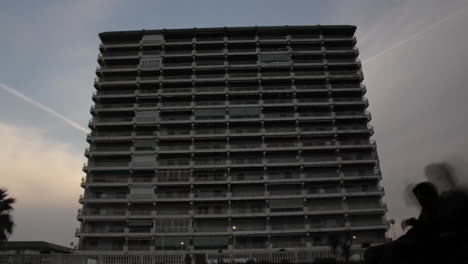 ghostlike figures drift past a massive apartment block at dusk, blending memory and reality—evoking nostalgia, presence, and the layers of urban life