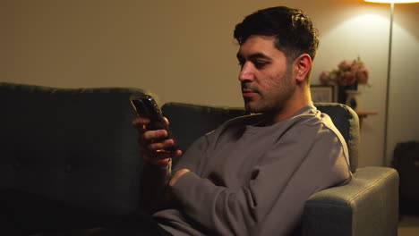 young man spending evening at home sitting on sofa with mobile phone scrolling through internet or social media 10