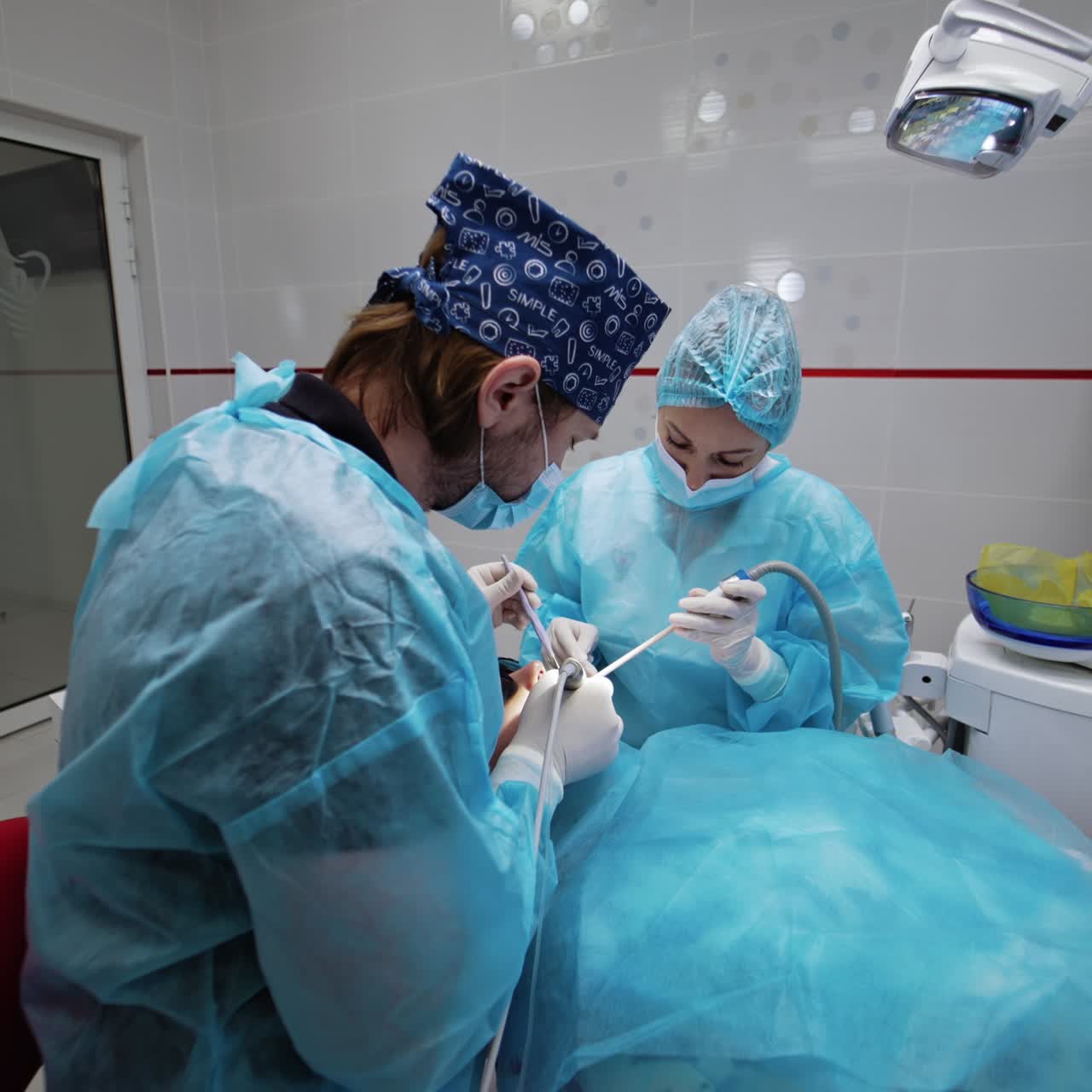 Bearded dentist holding instruments in both hands using them in treatment. Nurse sitting beside applies device
