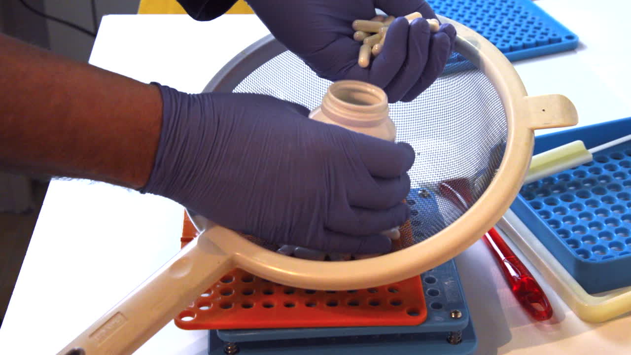 Pharmacist's hands with blue gloves sorting medical capsules into a bottle with professional equipment