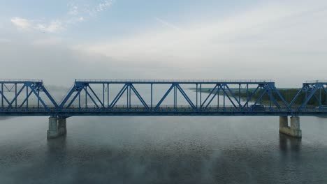 Vista-Aérea-Del-Puente-De-Acero-Sobre-El-Río-Lielupe-En-Una-Soleada-Mañana-De-Verano,-Niebla-Elevándose-Sobre-El-Río,-Autos-Conduciendo,-Gran-Drone-Dolly-Moviéndose-Hacia-La-Izquierda-Y-Baja