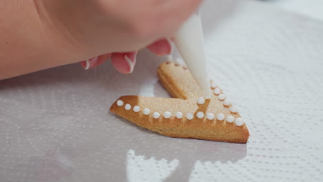 close-up of hand decorating heart-shaped cookie with white icing using a piping bag, intricate icing dots carefully placed on golden brown cookie resting on textured paper towel under soft lighting