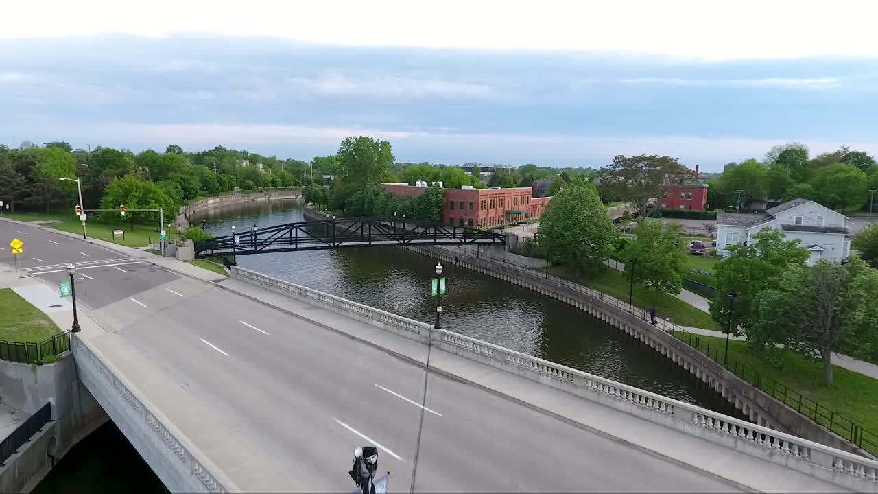 A drone flies over the Flint River and rises above an auto bridge and pedestrian bridge in downtown Flint, Michigan at dusk in summer.