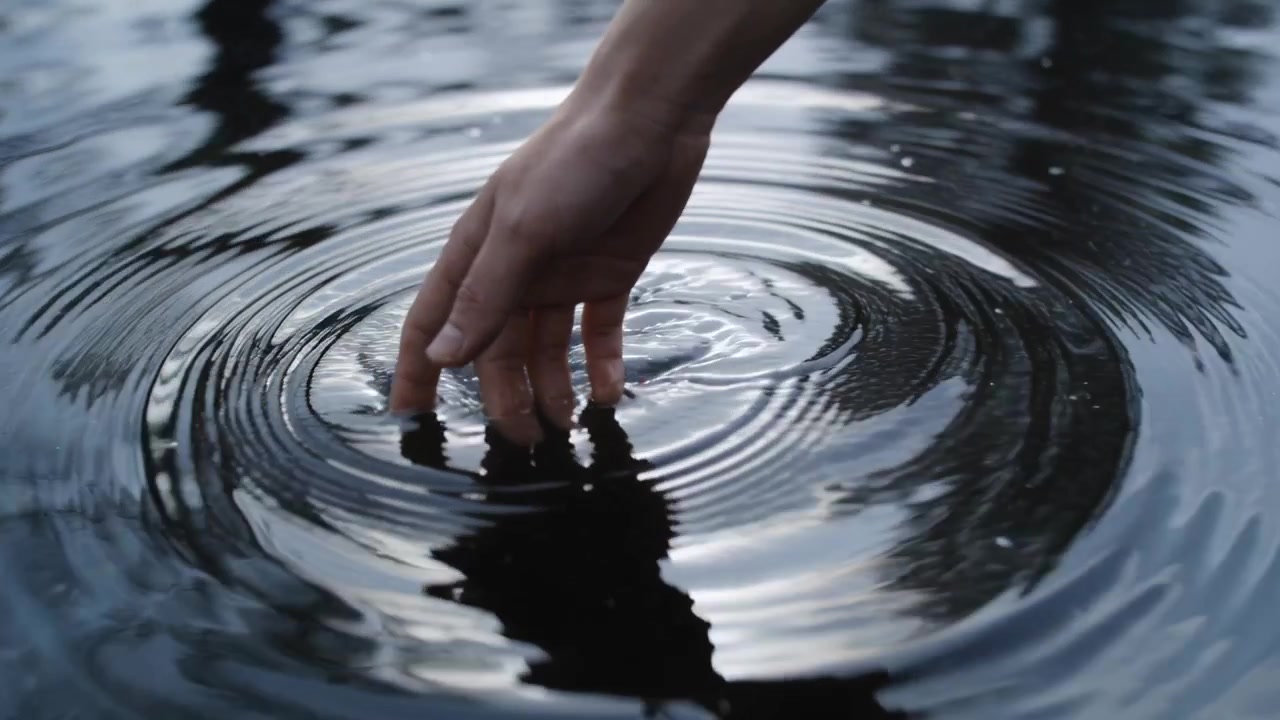 Punte delle dita di una mano nuda che premono toccando la superficie di uno stagno, creando increspature e distorcendo i riflessi degli alberi.