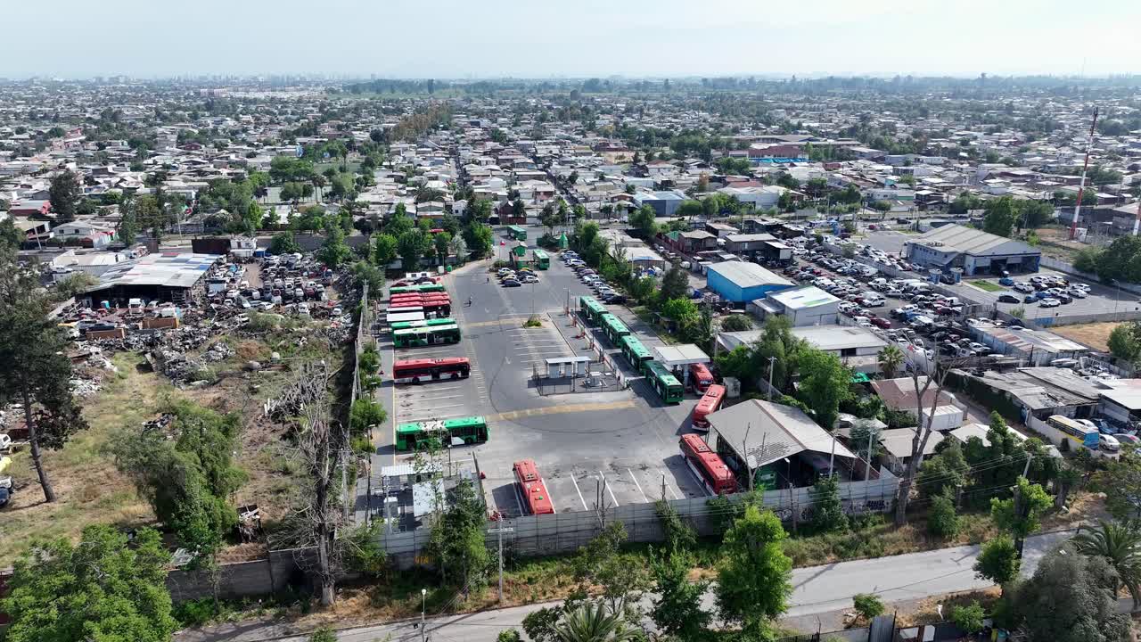 Forward drone aerial of bus depot terminal with green and red buses, parking lot, and urban residential neighborhood backdrop