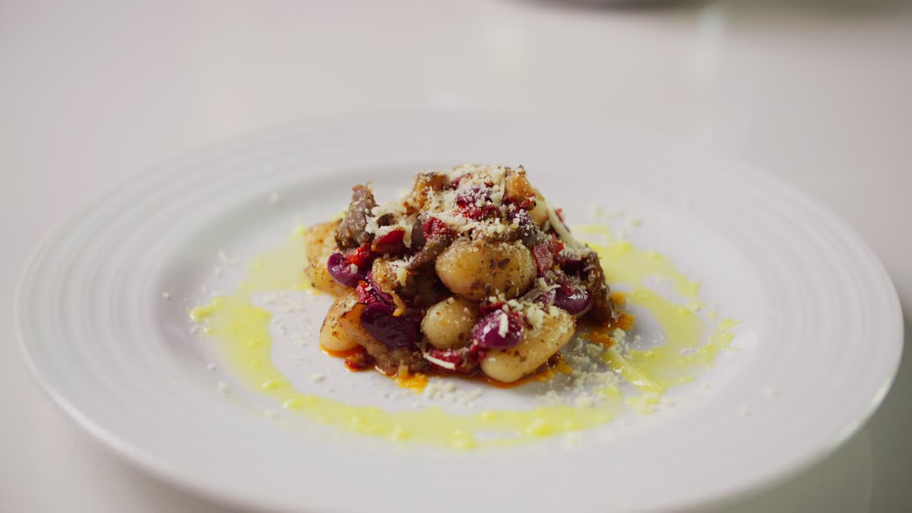 A white plate holds a gourmet dish of gnocchi topped with meat, red onions, tomato, grated cheese, and olive oil, artfully arranged with a drizzle of sauce around the rim.