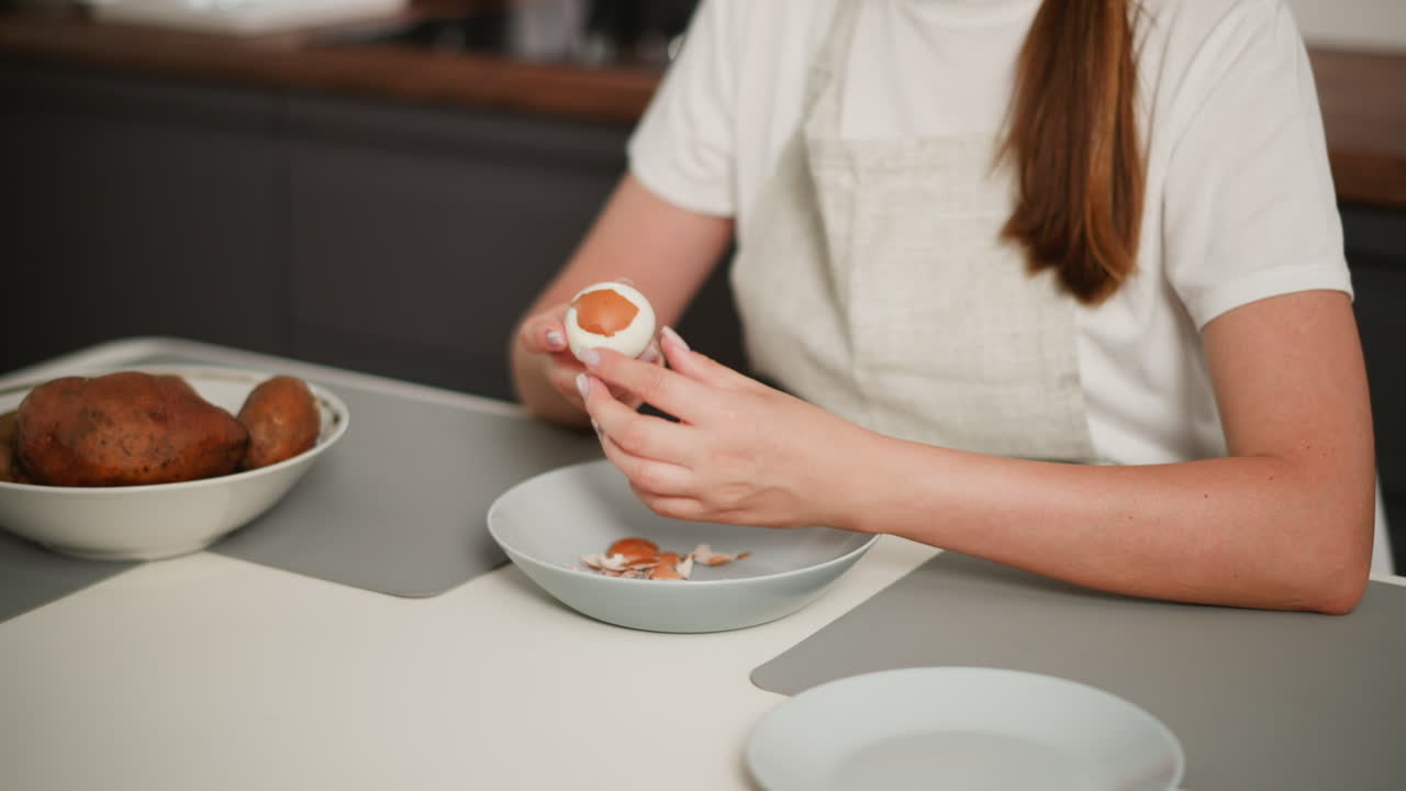 Partial view of girl in apron holding boiled egg while peeling off remaining shell over dish filled with fragments, seated at kitchen table with bowl of potatoes and plates on placemats in clean home