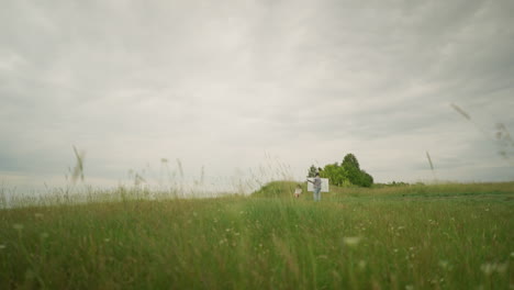 a back view of an artist, wearing a hat and checkered shirt, deeply focused on painting at an easel in a lush, green field under a cloudy sky. in front of him, a woman is seated