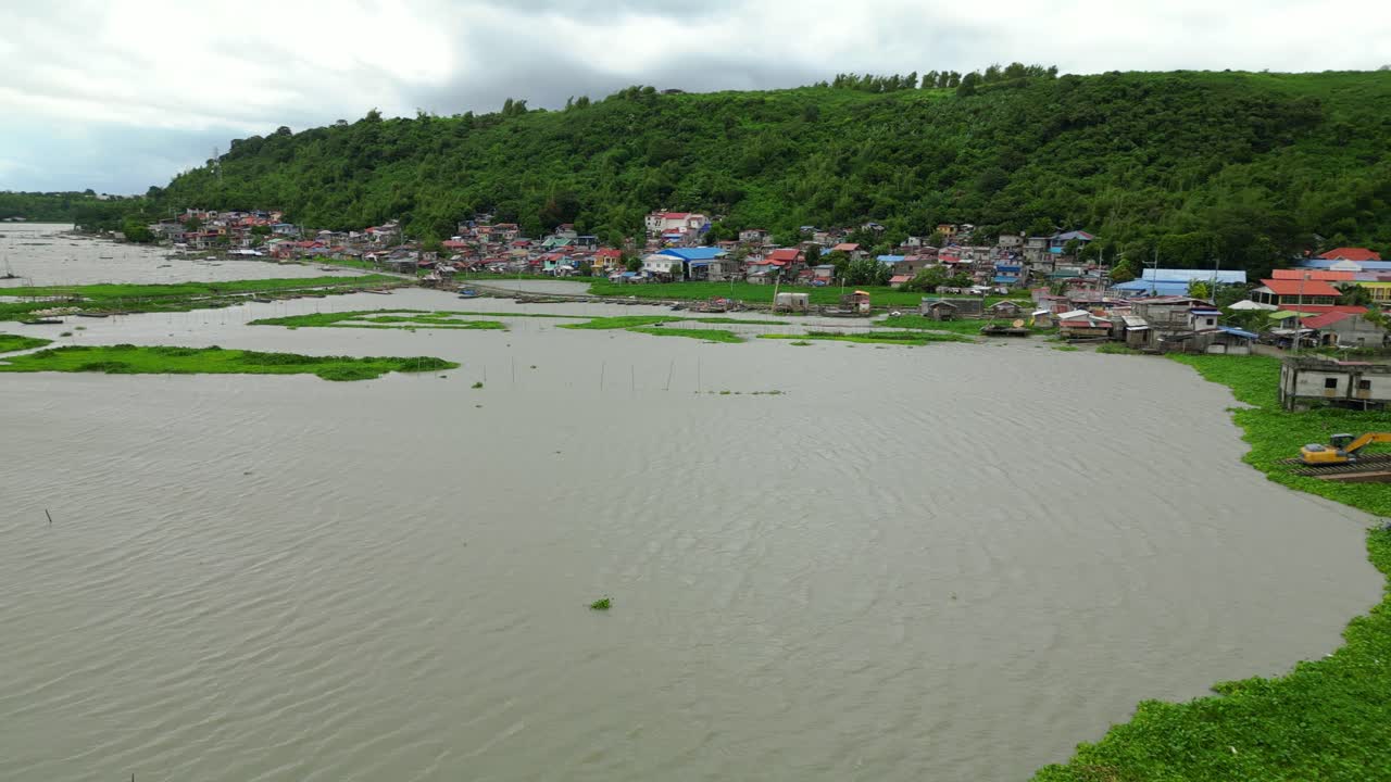 Drone pans over a shoreline village with vibrant rooftops and calm water, framed by lush hills and gray skies
