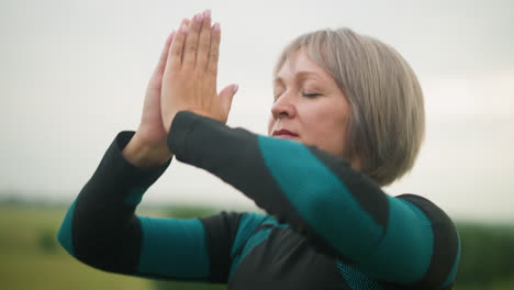 close up of middle-aged woman with arms raised, hands brought together in a meditative pose, practicing mindfulness outdoors in a misty grassy field under a cloudy sky