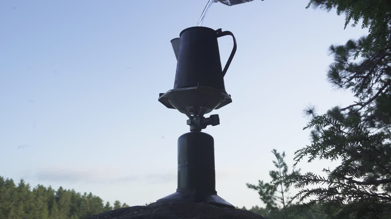 Pouring water into stove in wilderness for coffee preparation. Surrounded by forest