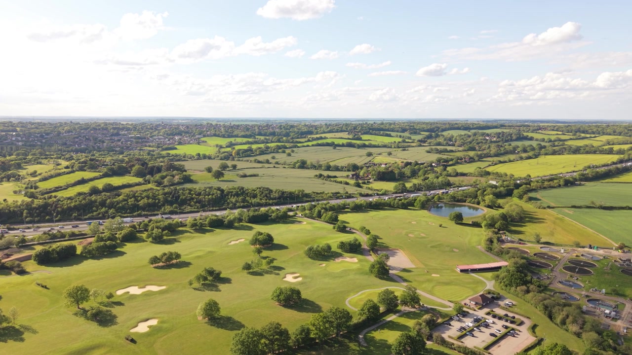 Drone flies over a verdant golf course adjacent to a busy highway. Vast green fields and scattered trees blanket the rural area beneath a bright blue sky filled with white clouds.