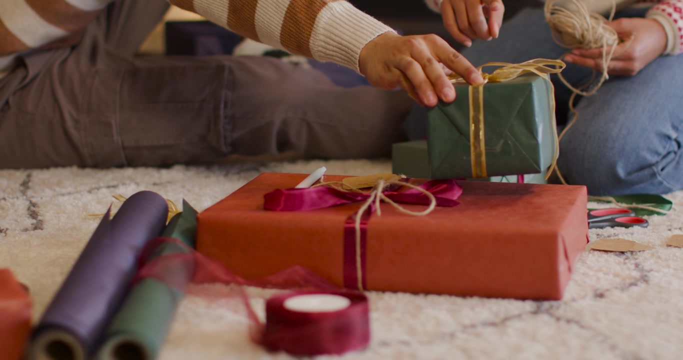 Pareja afroamericana con suéteres, preparándose para las fiestas, envolviendo un regalo rojo y apilando una caja sobre la alfombra de casa.