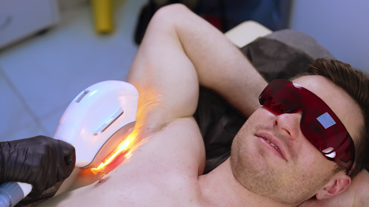 Man in goggles lying with hands under the head. Apparatus for laser epilation is moved by the armpit zone. Close up.