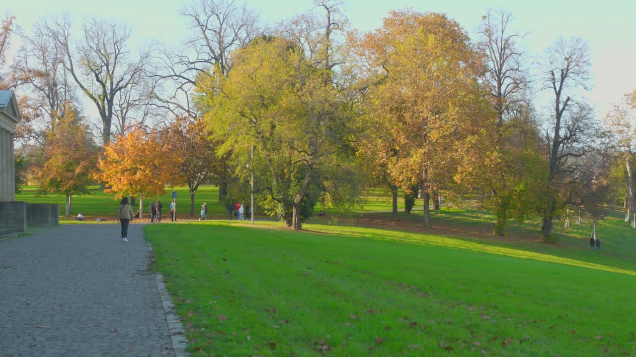 People Walking At The Schlossgarten Park With Autumnal Trees On A Sunny Day In Stuttgart, Germany. - wide shot