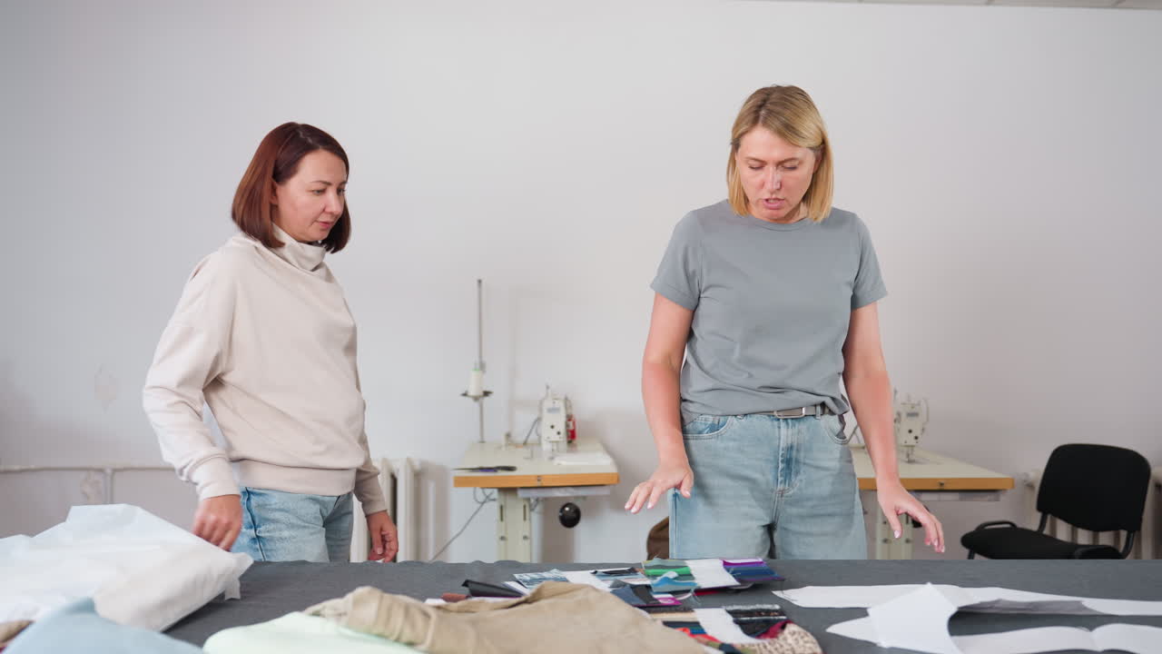 Wide view of tailor shop as two women walk in, one explaining different color combinations to colleague while standing near table filled with colorful fabrics, sewing machines and workspace tools around
