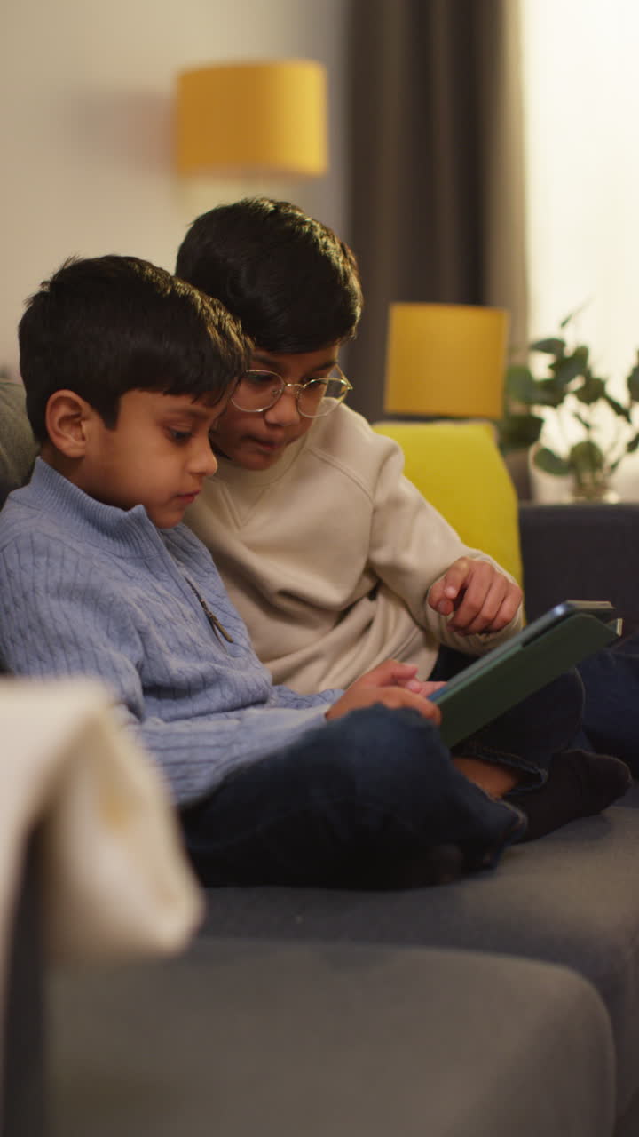 Vertical Video Of Two Young Boys Sitting On Sofa At Home Playing Games Or Streaming Onto Digital Tablet Together 1