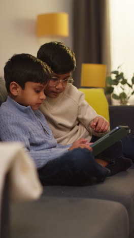 Video-Vertical-De-Dos-Niños-Sentados-En-Un-Sofá-En-Casa-Jugando-O-Transmitiendo-Juntos-A-Una-Tableta-Digital-1