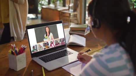 Schoolgirl-using-laptop-for-online-lesson-at-home,-with-diverse-teacher-and-class-on-screen