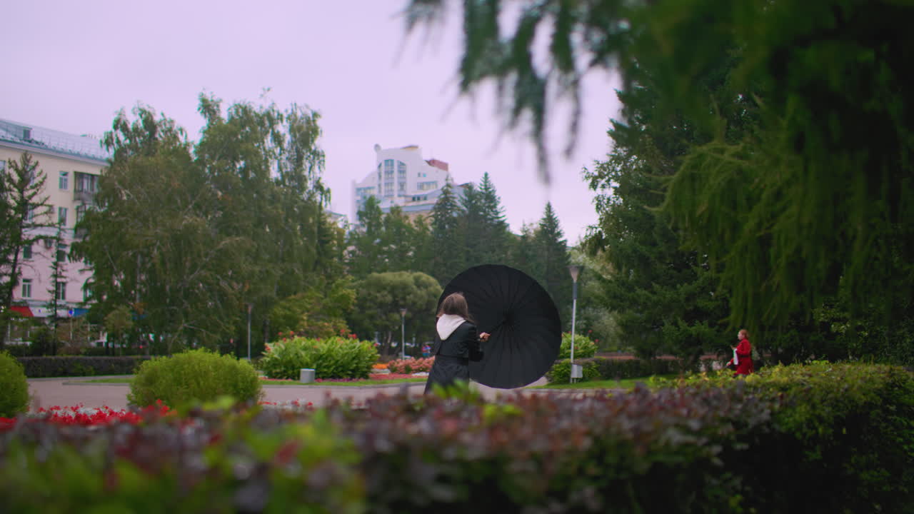 Pretty woman in black coat holding umbrella walking through park with green trees, bushes and flowerbeds on windy day with urban buildings in background creating moody seasonal city landscape atmosphere