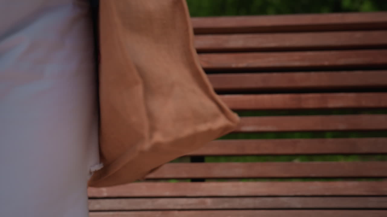 Close up rear view of woman in blue shirt and white pants carrying beige tote walking toward wooden bench and sitting down under sunlight in peaceful park