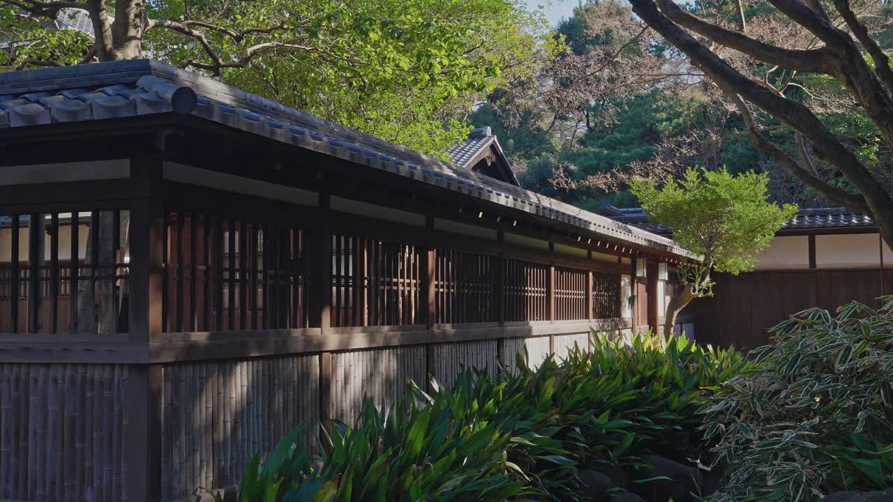 日本建築の木製外壁と格子をスムーズに右パンし、鮮やかな緑の植物と静かで日当たりの良い庭の雰囲気を映し出す。