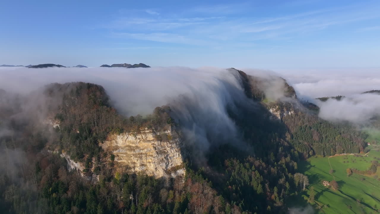 Thick fog cascades over a cliffside in the Jura mountains on a sunny day creating a dramatic view