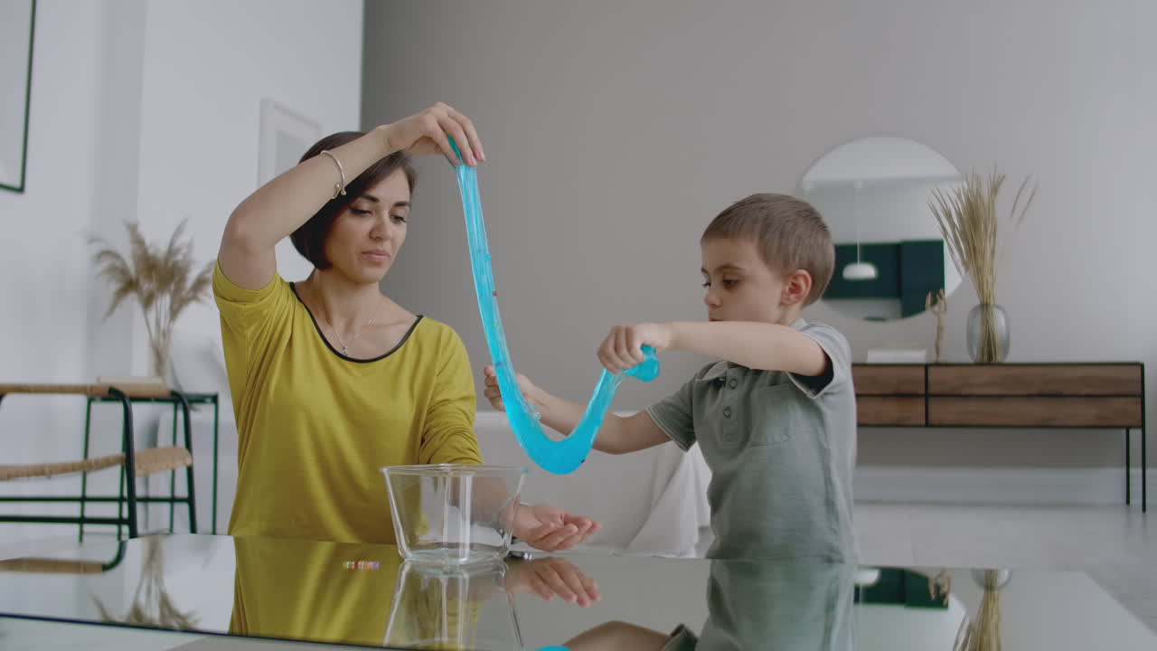In a white apartment sitting on the floor mom and boy used to play a stretchable plasticine toy laughing and smiling together.