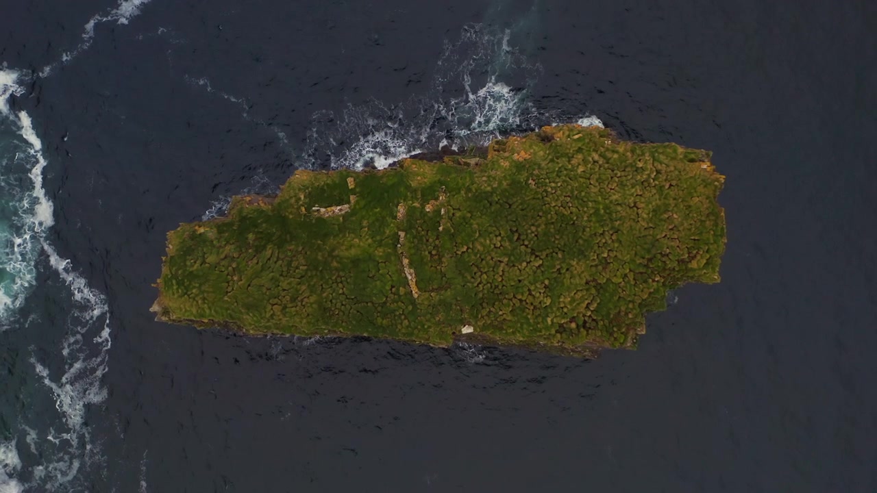 Drone subindo em vista superior sobre o farilhão Dun Briste, rodeado por ondas e água branca agitada em oceano azul profundo.