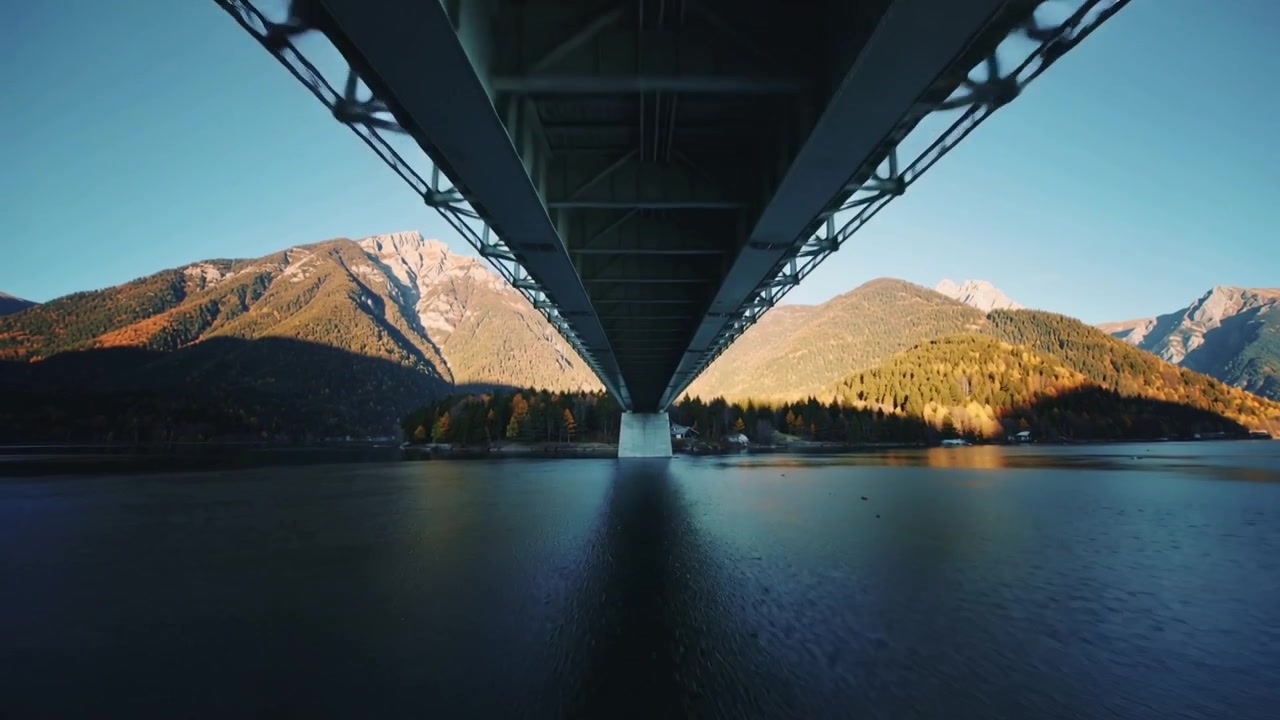 Telecamera che si allontana rivelando la parte inferiore di un ponte d'acciaio su un lago di montagna, con il riflesso del molo