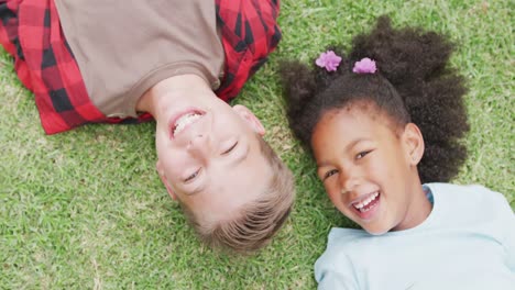 Overhead-video-portrait-of-smiling,-diverse-boy-and-girl-lying-on-grass-in-schoolyard,-copy-space