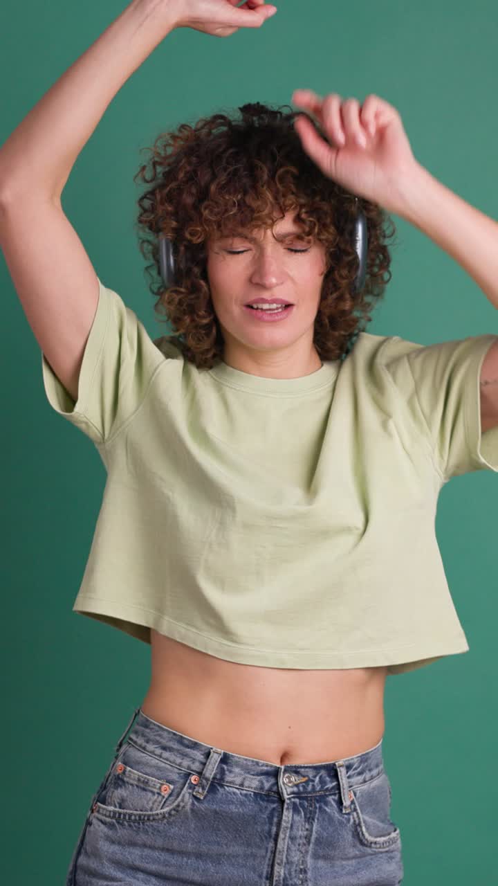 Adult woman dancing with headphones, green studio background. Vertical footage