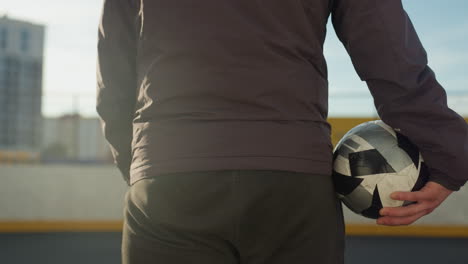 vista trasera de primer plano de un individuo sosteniendo una pelota de fútbol, listo para entrar en la arena deportiva, mostrando ropa atlética y telón de fondo urbano iluminado por la suave luz del sol