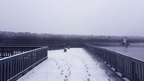 Volando-Sobre-Un-Embarcadero-Cubierto-De-Nieve-En-El-Lago-Strzeszynskie-Y-Mirando-El-Agua-Oscura,-El-Aireador-Y-Los-árboles-Helados-En-La-Otra-Orilla