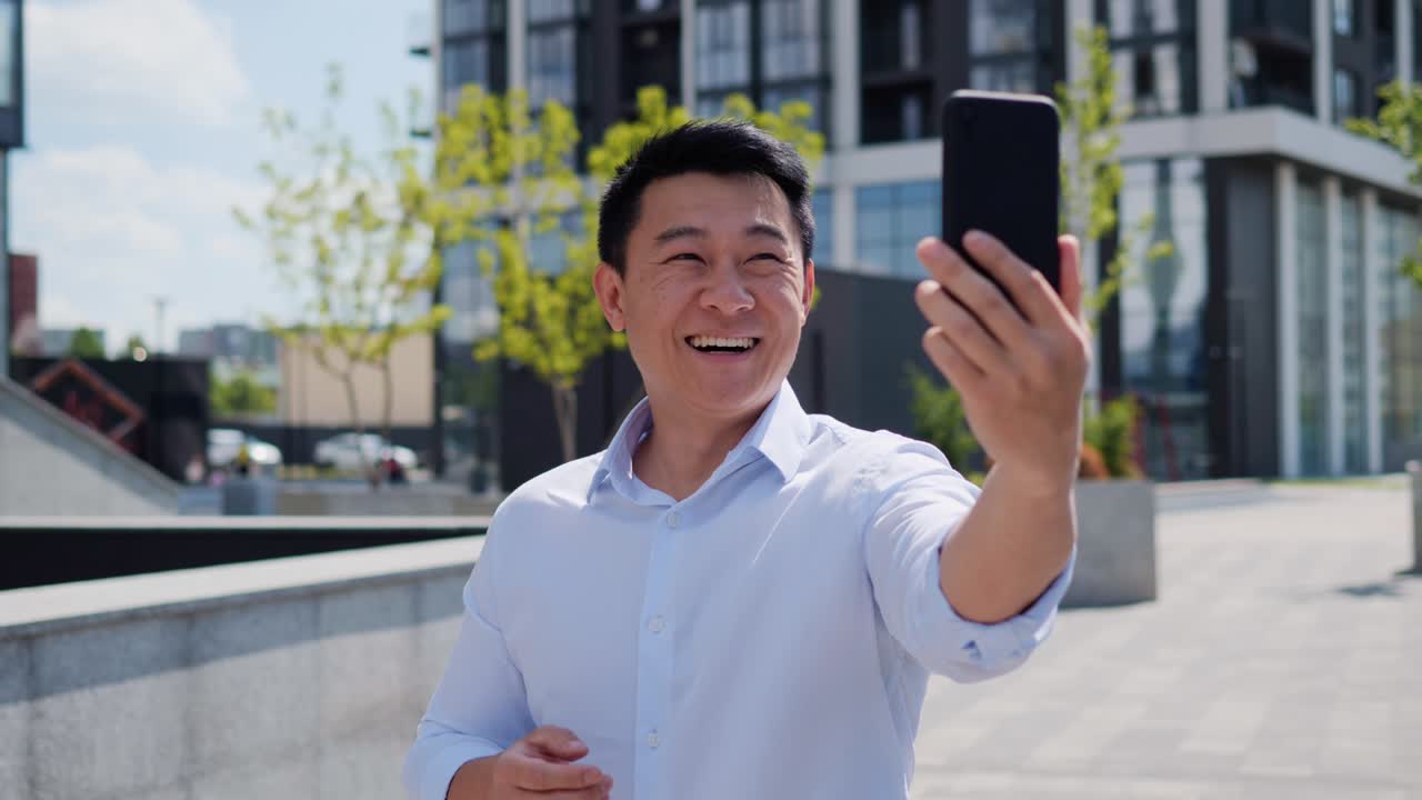 일본인 사업가가가 온라인 영상통화를 하고 손을 흔들며 휴대전화를 이용해 미소 짓고 있는 모습, 멋진 아시아 남성이 비즈니스 디스트릭트 밖에서 서 있는 모습, 사업가 블로거의 초상화 촬영 영상.