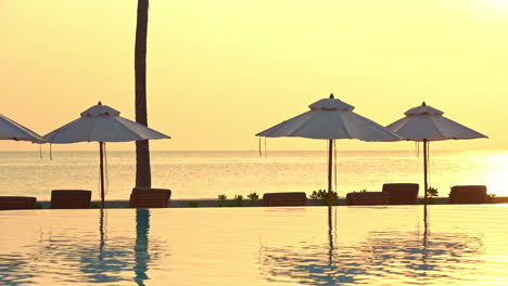 a lovely view of a romantic sunset on a clear golden sky and calm ocean in the background with empty chairs, umbrellas and a swimming pool in the foreground