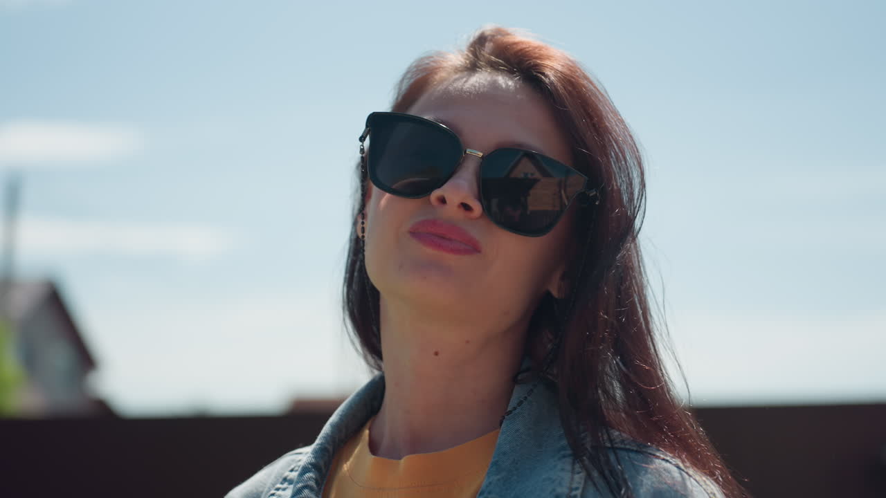 Young woman with long dark hair wearing black sunglasses, orange shirt, and denim jacket poses confidently outdoors under clear sunny sky, with blurred house and poles in background