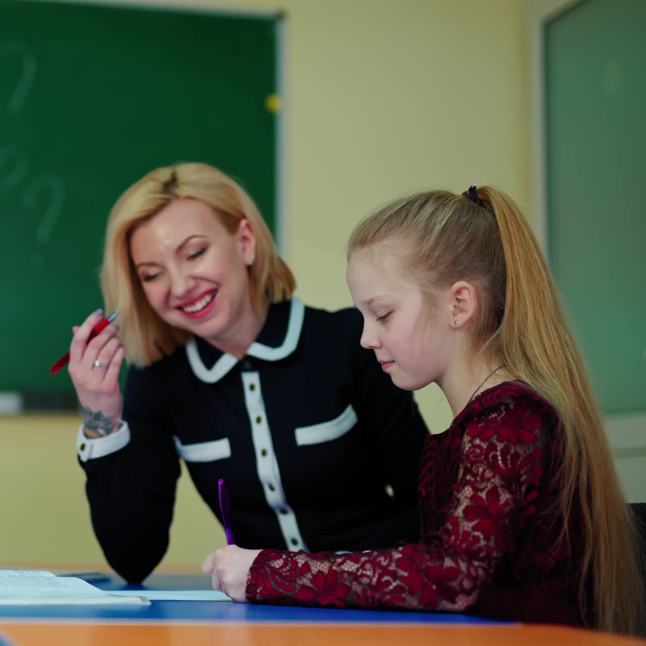 Teacher helps schoolgirl. Little girl and her teacher sitting in the classroom. Woman teaching a clever student on the blackboard background at school.