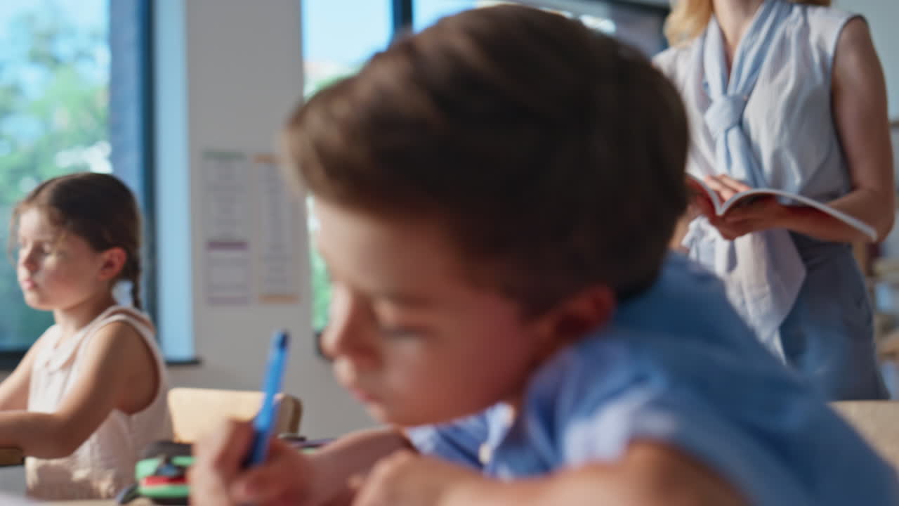 School teacher walking classroom with book observing studying pupils writing