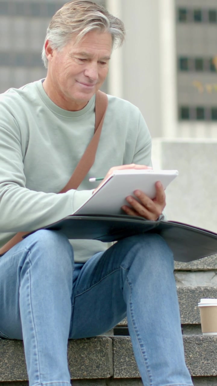 Vertical video: Sitting on steps man in green sweater sketching with pencil on pad, cup nearby
