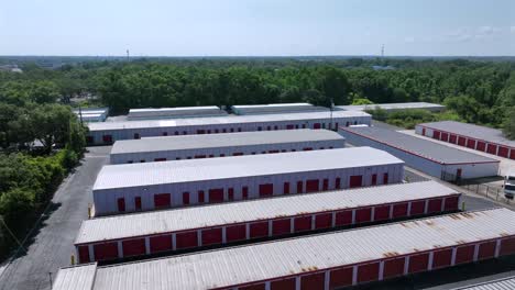 green self storage unit buildings or warehouse temporary storage at sunny day in florida. aerial approaching shot. american suburb garage with forest trees in background.