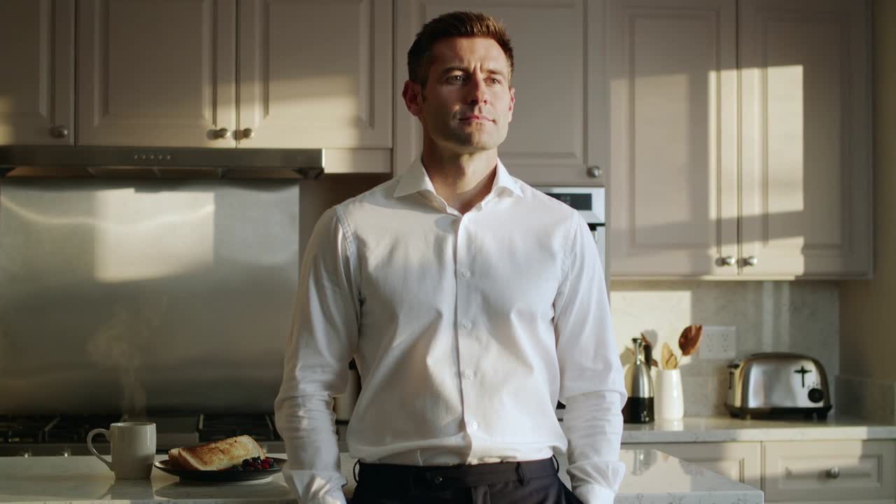 Ein Mann in weißem Hemd und dunkler Hose blickt kurz auf, während er sich mit Tasse und Toast an der Küchentheke verlagert.