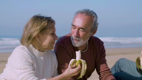 senior man and woman sitting on sandy shore and eating bananas