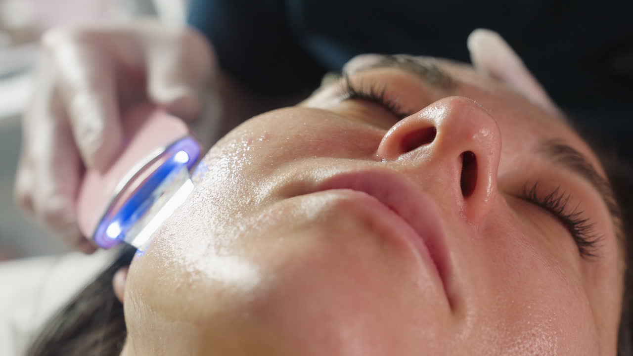 Close up of visitor closing eyes peacefully face up as technician glides steel head massager in circular motions along side jawline during spa session for soothing facial therapy under soft lighting