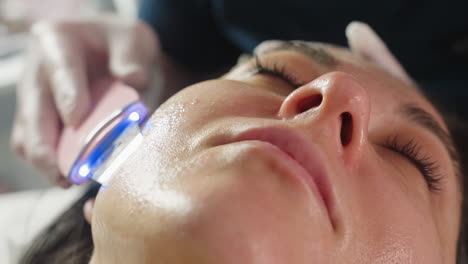 close up of visitor closing eyes peacefully face up as technician glides steel head massager in circular motions along side jawline during spa session for soothing facial therapy under soft lighting