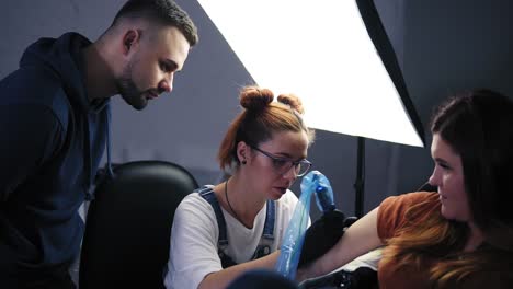 the girl - the master of tattoos providing a lesson for man standing above and watching the process. a girl holding a tattoo machine and applied in a pattern on the skin of her female client