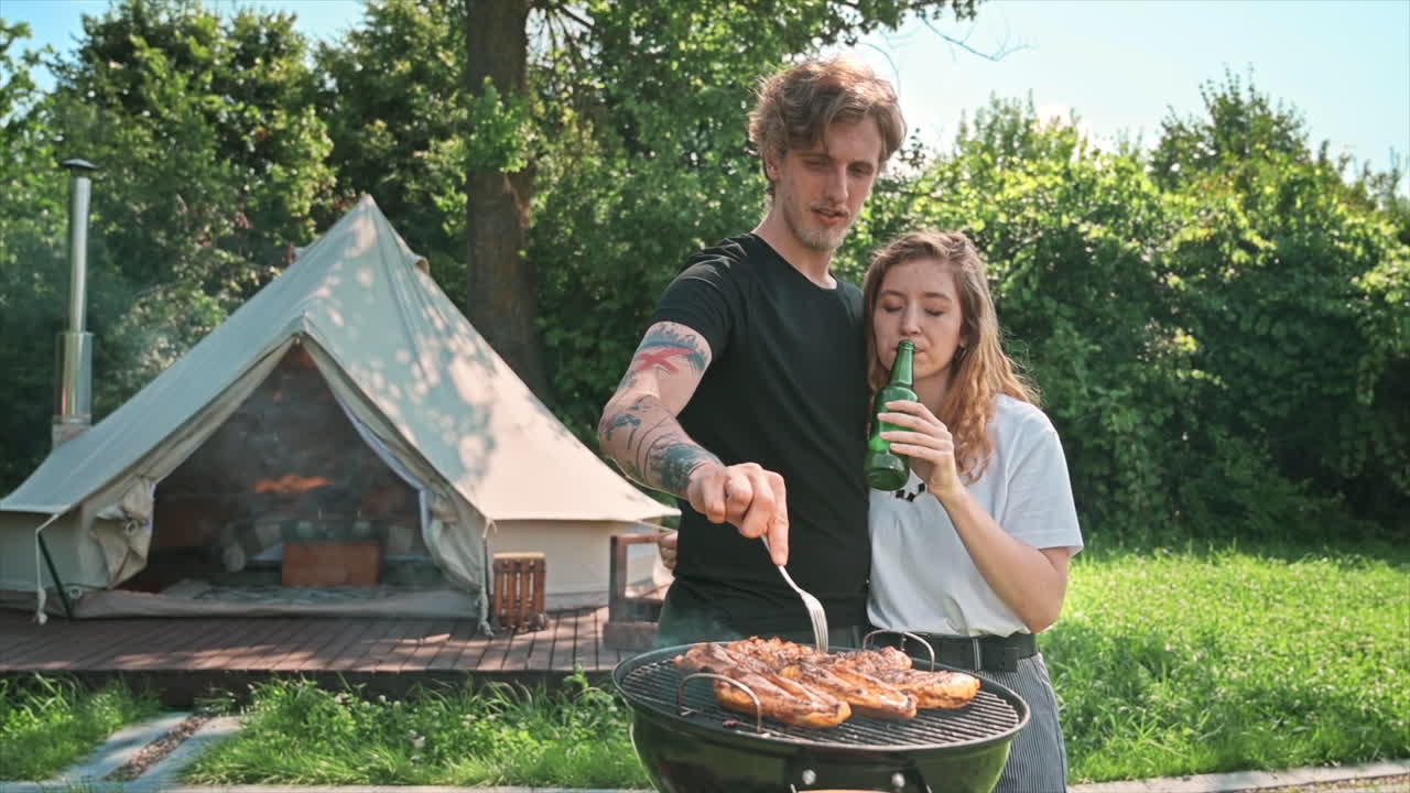 Giovane coppia che frigge carne alla griglia con birra. Vegetazione circostante. Tenda da glamping sullo sfondo. Slow motion