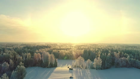 Drone-flight---snow-covered-clearing-with-remote-cabin,-yellow-sunset-over-forested-landscape