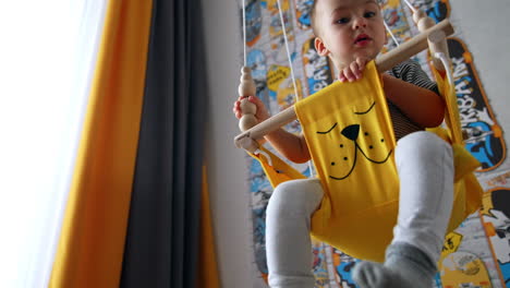 lovely little baby swinging in the swing in the room. kid plays with beads on the rope of a swing. low angle view.